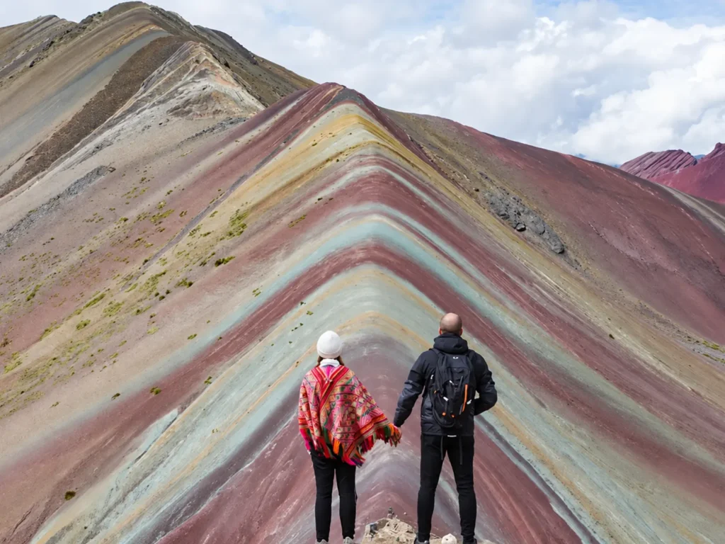Couple holding hands while facing the colorful mineral bands of Rainbow Mountain, Peru.
