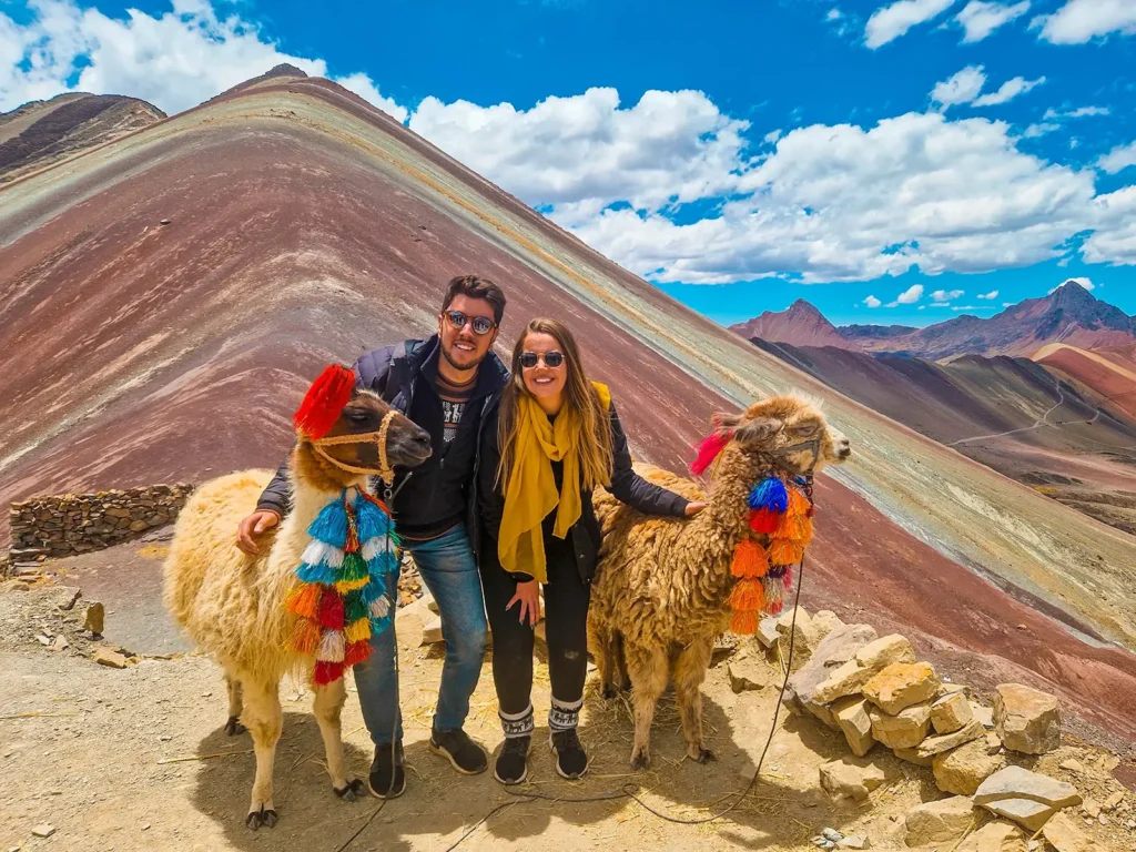 Couple posing with two decorated llamas beside the colorful slopes of Rainbow