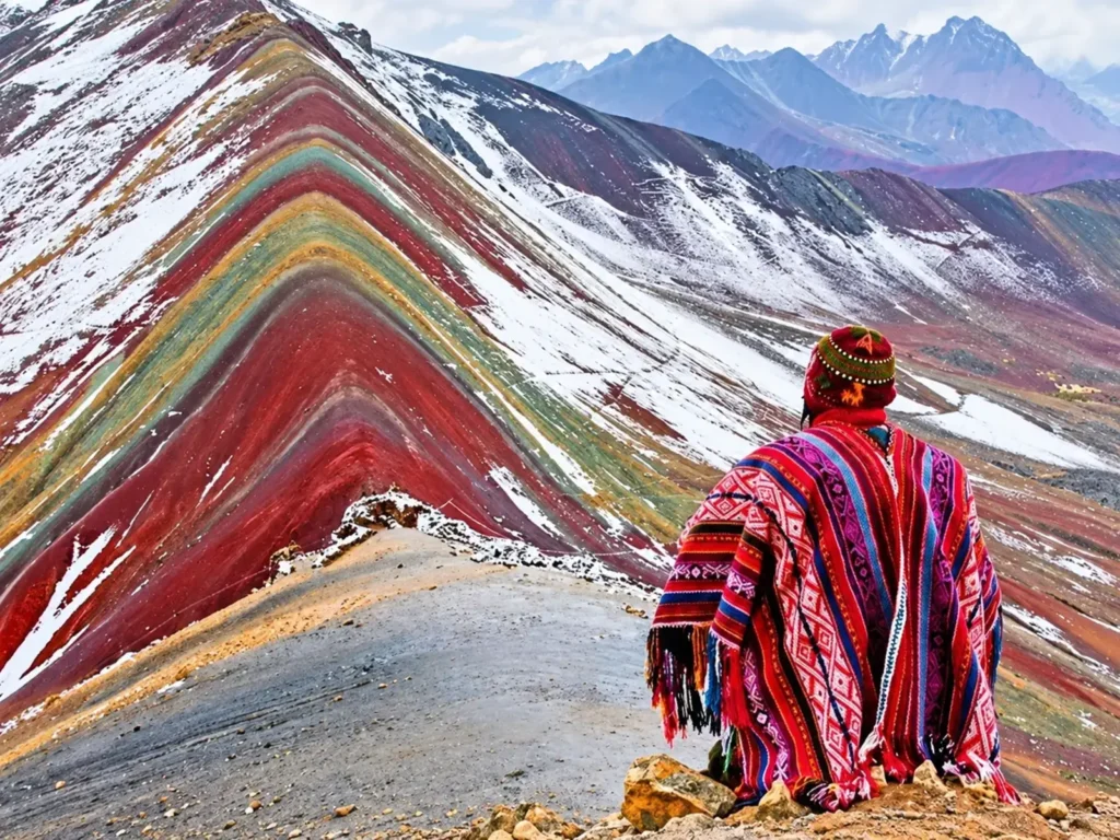 Traveler wearing a red Andean poncho overlooking the colorful, snow-dusted slopes of Rainbow Mountain.