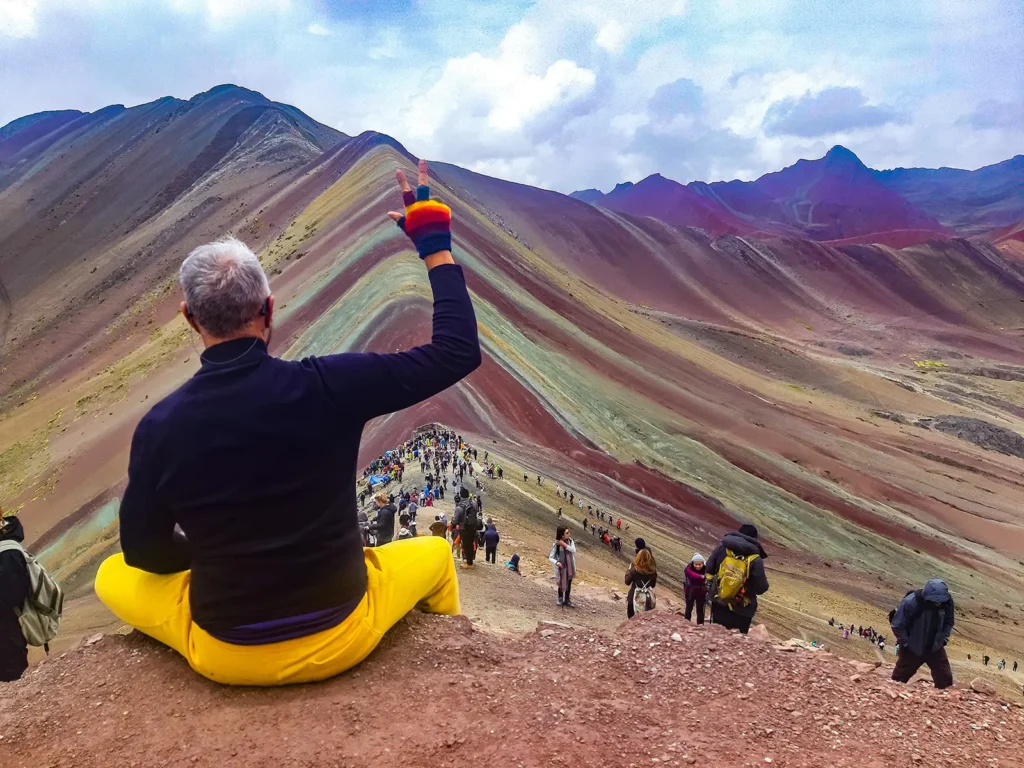 Traveler in yellow pants overlooking Rainbow Mountain and hikers at the viewpoint in Cusco, Peru.